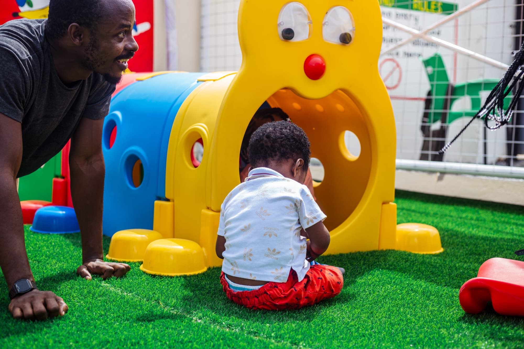 A toddler exploring a yellow play tunnel while his father watches